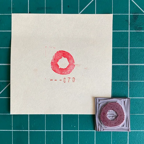 The composition features a cream paper with a red stamped ring and the numbers --070, next to a small square object displaying a reddish ring framed by layered white geometric shapes.