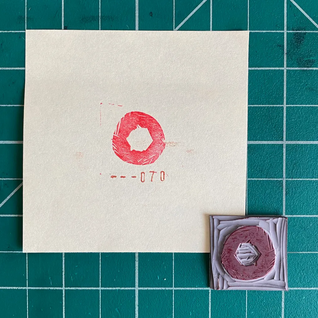 The composition features a cream paper with a red stamped ring and the numbers --070, next to a small square object displaying a reddish ring framed by layered white geometric shapes.