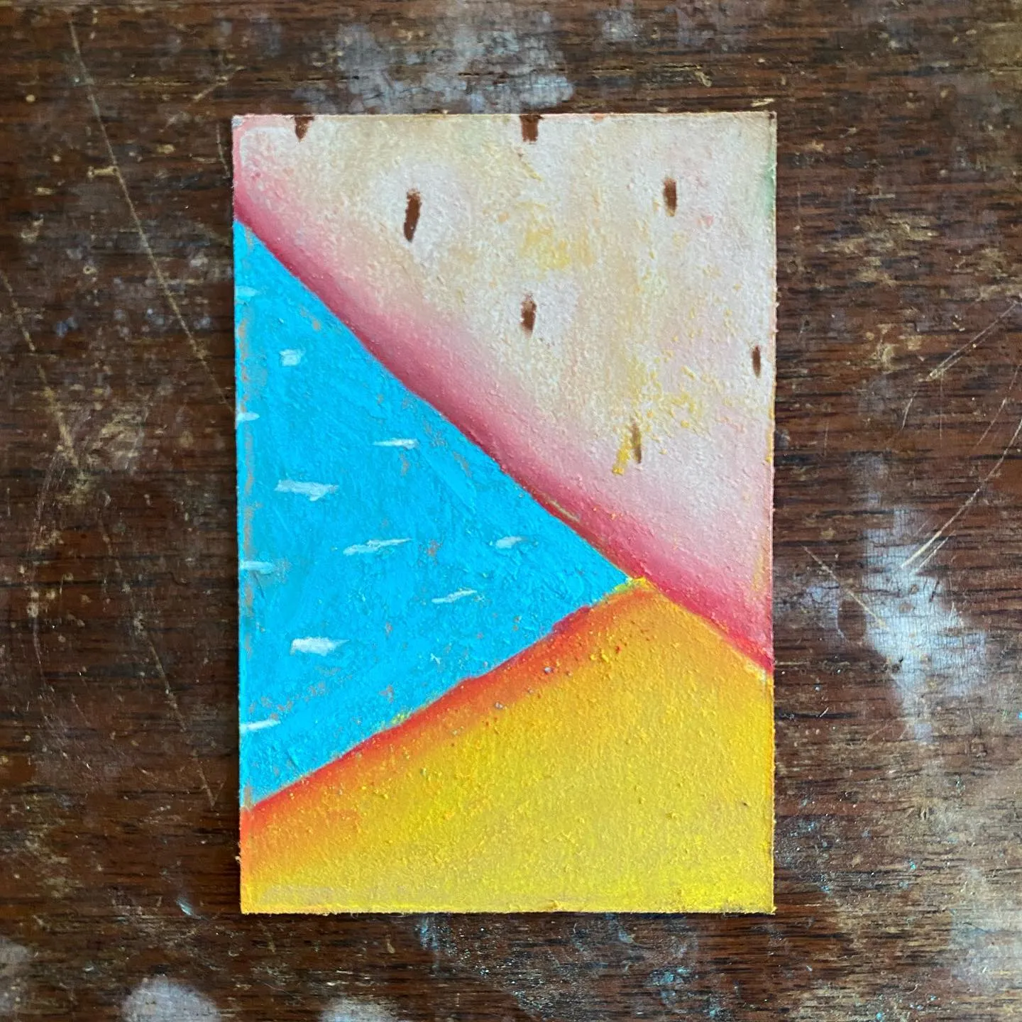 A square painting on a dark wood surface features a diagonal composition of bright turquoise blue, yellow-orange, and pale cream, separated by a muted reddish band.
