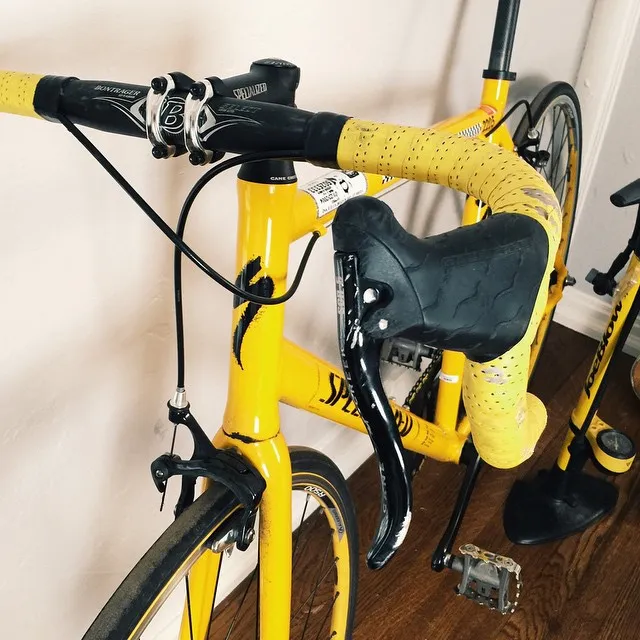 A bright yellow bicycle is angled on a dark floor, featuring textured yellow handlebar wraps and a matching seat, with black tires and cables visible.