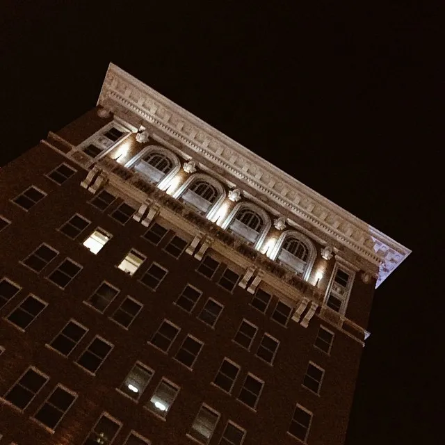 A sharply angled view of a multi-story brown building facade set against a dark sky, featuring repetitive windows and an ornate upper section with arched architectural details.