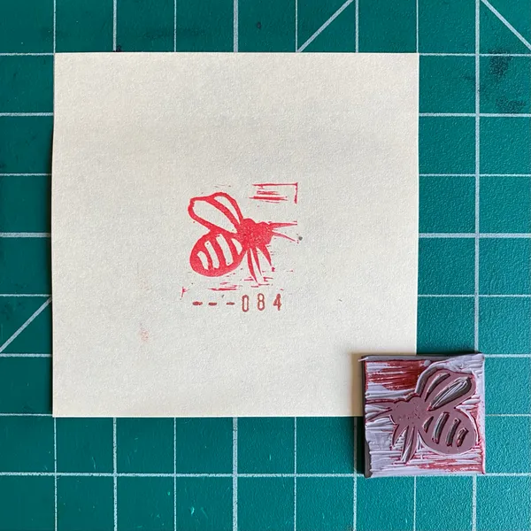 A cream square paper featuring a red stylized bee and the number 084 sits on a teal grid, next to a small reddish-brown square with a matching bee design.