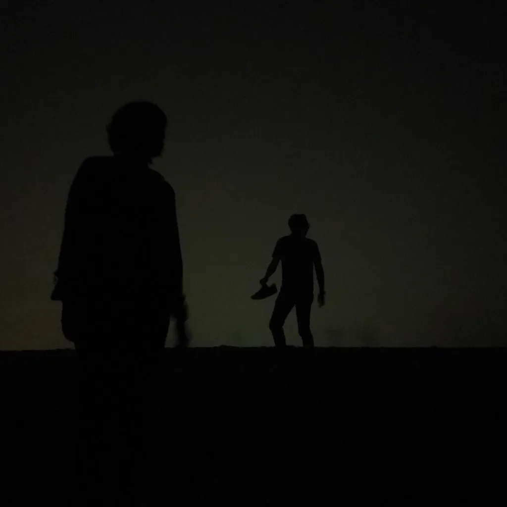 Two dark silhouettes of people stand on a horizon line against a muted, dark gradient sky.