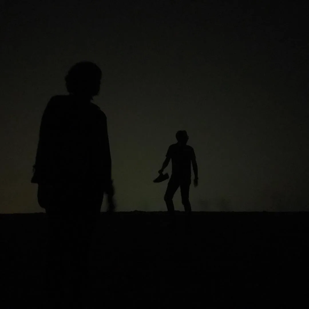 Two dark silhouettes of people stand on a horizon line against a muted, dark gradient sky.