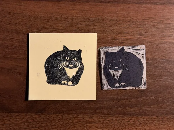 Two stylized black cat portraits rest on a dark wood background. The left cat is on a solid cream square, while the right cat is on a smaller, distressed rectangular piece.