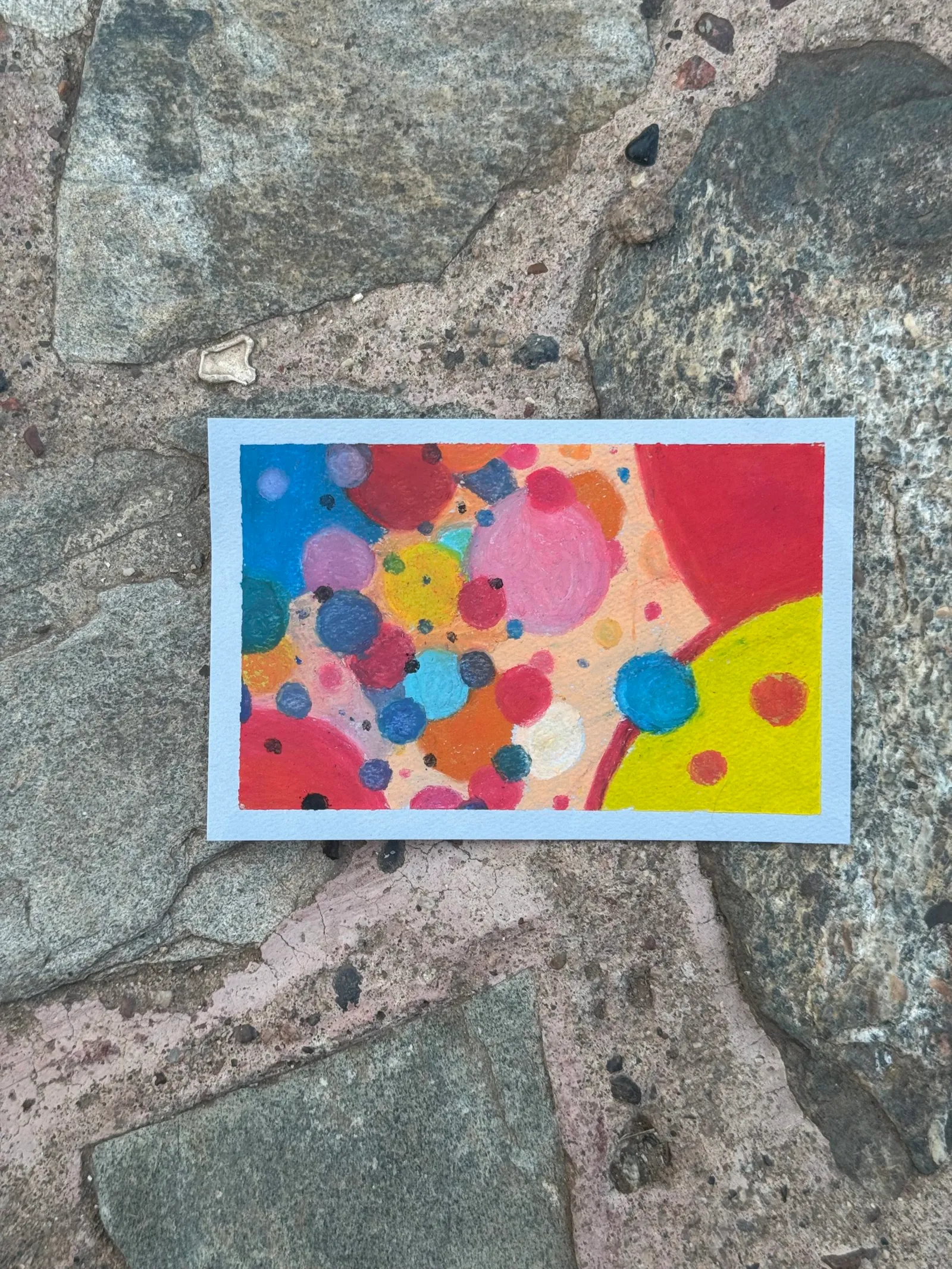 A vibrant abstract composition of overlapping circles and irregular shapes in bright reds, yellows, blues, and pinks, creating a dense, colorful pattern displayed on rough gray stones.