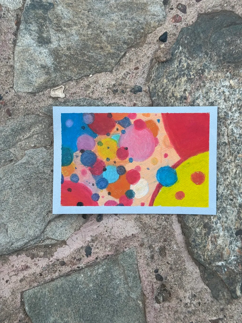 A vibrant abstract composition of overlapping circles and irregular shapes in bright reds, yellows, blues, and pinks, creating a dense, colorful pattern displayed on rough gray stones.