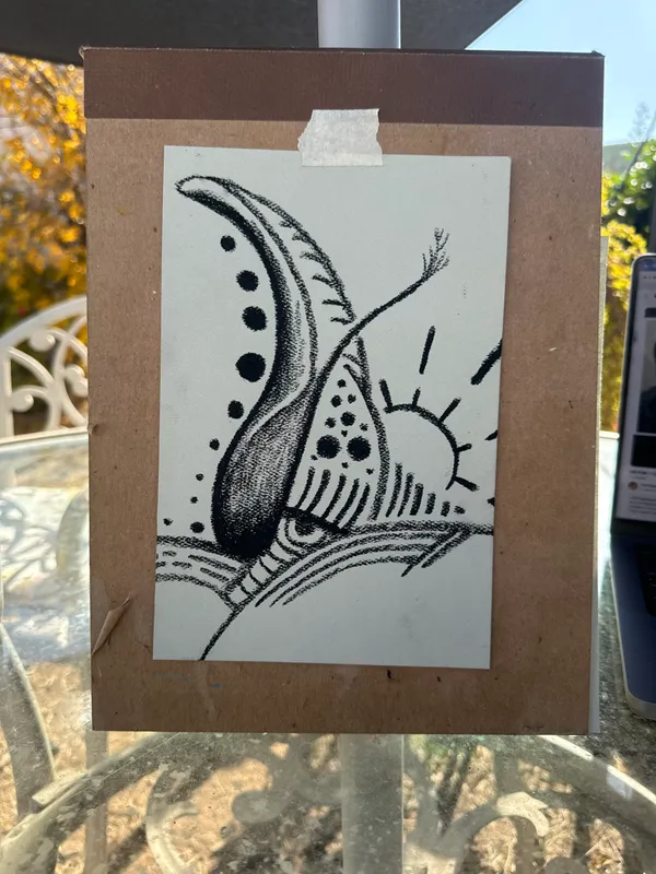 A black and white drawing features a stylized, curved creature with internal dots and patterns, rising from horizontal lines on white paper, taped to a brown cardboard box.