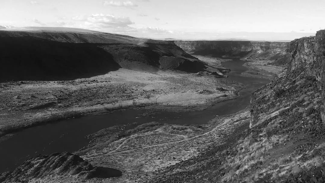 High contrast black and white photograph of a winding river cutting through a deep canyon, flanked by dark, textured cliffs and distant rolling hills under a bright sky.