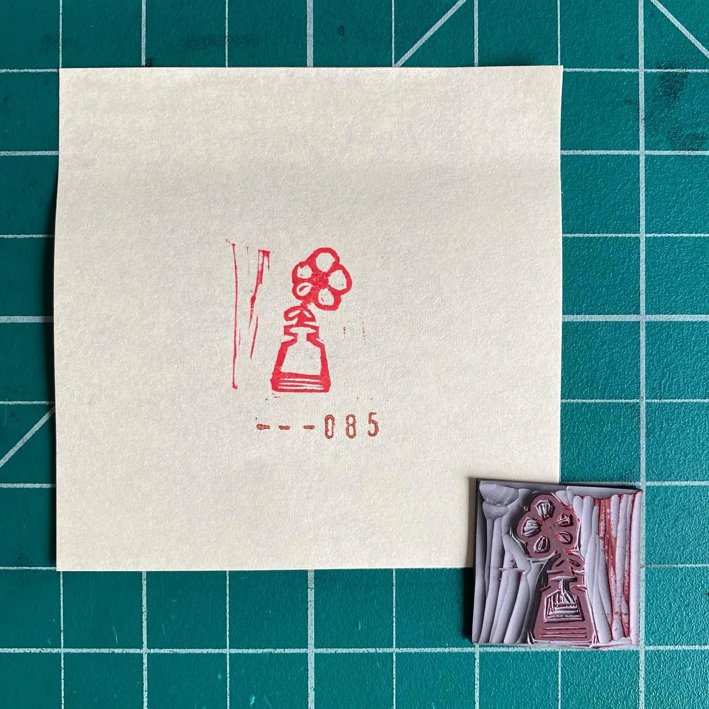 On a teal grid, a cream paper displays a red line drawing of a bottle and flowers, marked --085, next to a small, textured red and white square featuring a stylized object.
