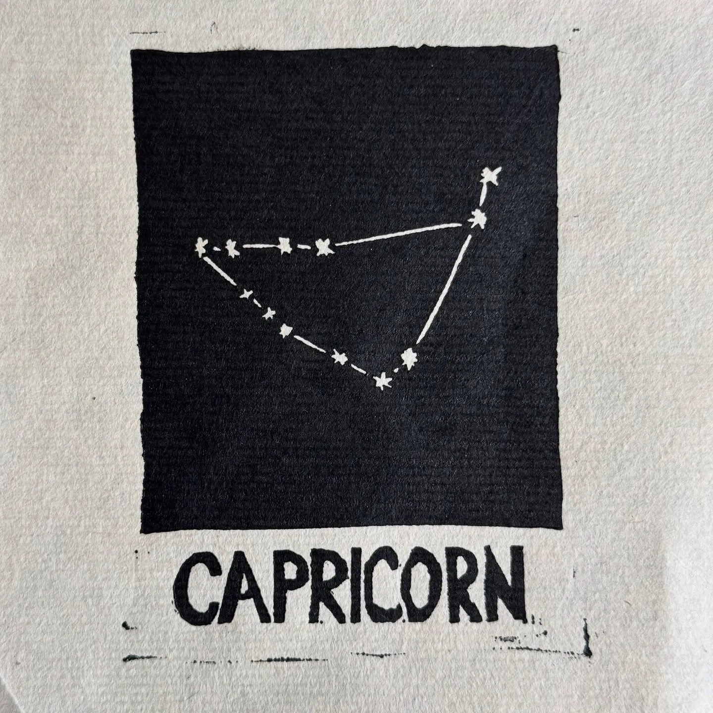 A black square on light fabric features a white line drawing connecting stars in a constellation pattern, with the word CAPRICORN printed in large block letters beneath the square.