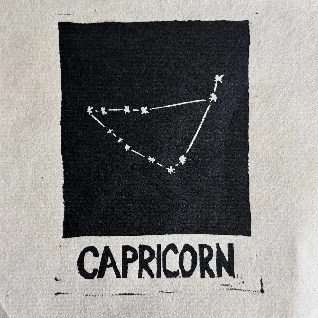 A black square on light fabric features a white line drawing connecting stars in a constellation pattern, with the word CAPRICORN printed in large block letters beneath the square.