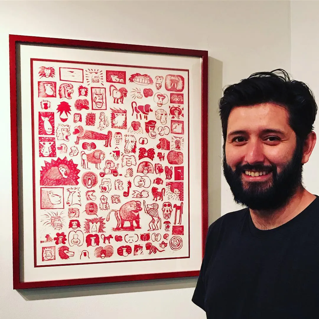 A man smiles next to a large, framed artwork filled with hundreds of small, stylized red drawings of figures and objects arranged in a dense grid on white paper.