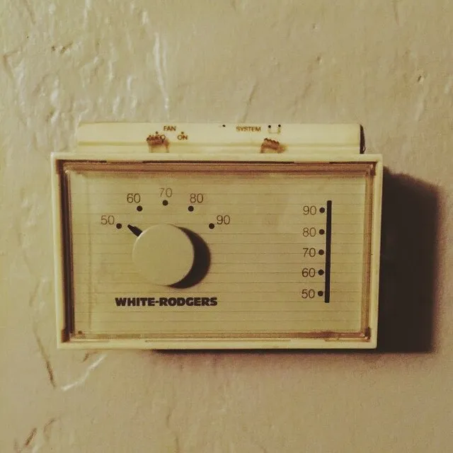 A cream-colored, rectangular control panel labeled WHITE-RODGERS is mounted on a textured beige wall, featuring a dial marked 50 to 90 and two switches set to ON.