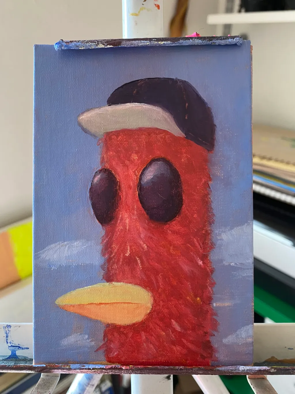 A stylized red creature with large black eyes and a yellow beak stands against a light blue background featuring white cloud strokes, wearing a black hat with a white band.