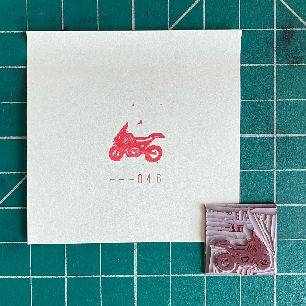 On green graph paper, a white square features a red graphic of a motorcycle and the number --046, positioned above a textured, reddish relief piece of a motorcycle.