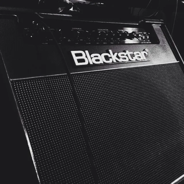 The front panel of a black amplifier dominates the frame, featuring the visible brand name Blackstar, a speaker grille with a dotted texture, and various control knobs under dramatic lighting.
