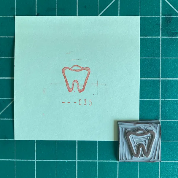 A light mint square rests on a dark green grid, featuring a red outline drawing of a tooth and the number --035, next to a small, sculpted brown object replicating a tooth structure.