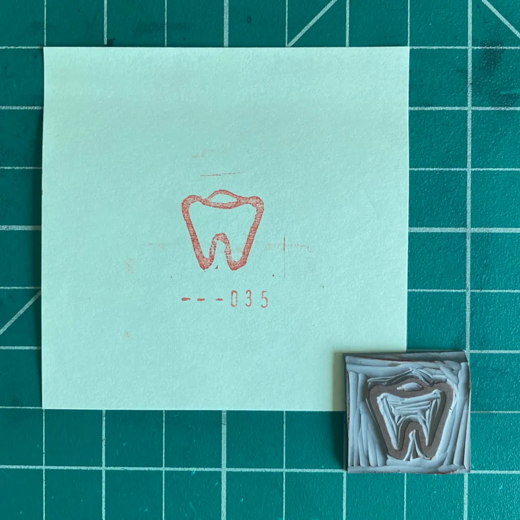 A light mint square rests on a dark green grid, featuring a red outline drawing of a tooth and the number --035, next to a small, sculpted brown object replicating a tooth structure.