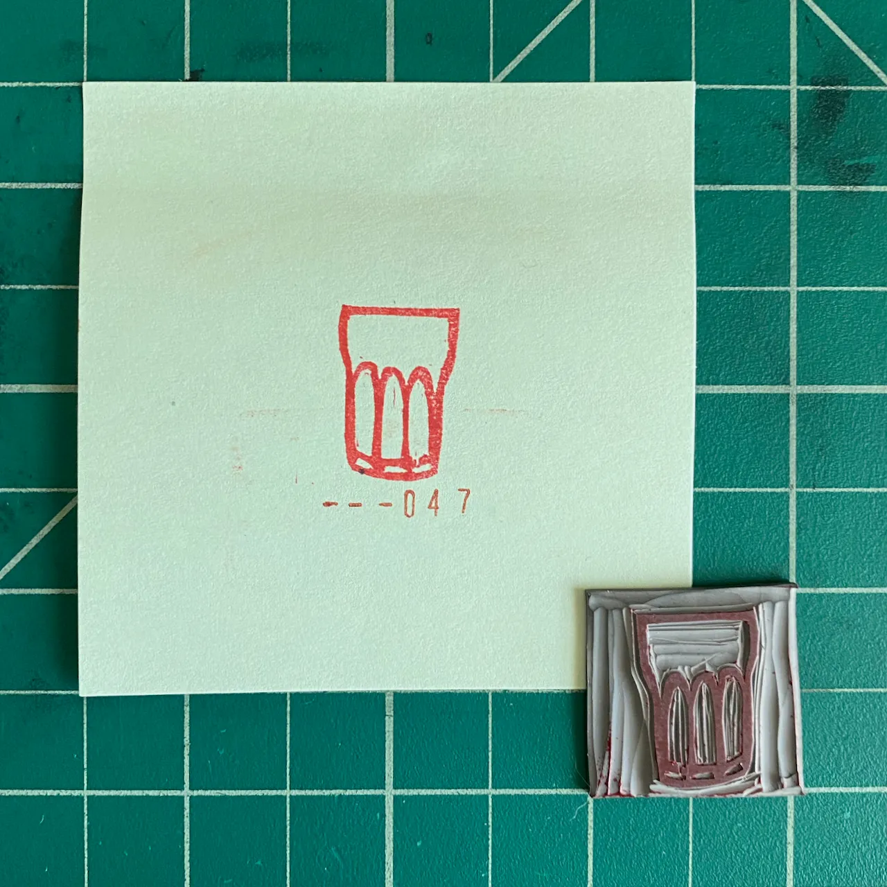The composition features a pale note on a dark grid background, displaying a red outline drawing of a glass and --047, alongside a small, rectangular, textured object of brown and gray tones.