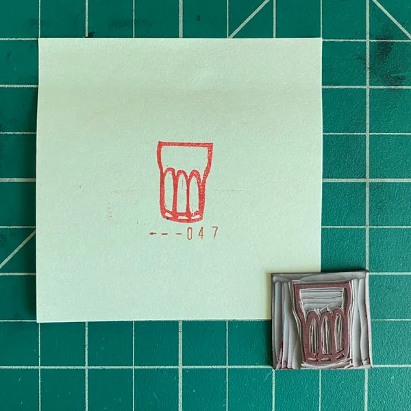 The composition features a pale note on a dark grid background, displaying a red outline drawing of a glass and --047, alongside a small, rectangular, textured object of brown and gray tones.