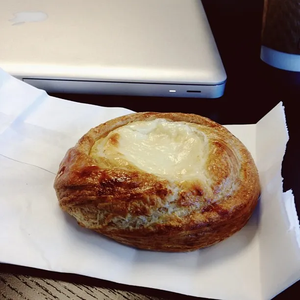 A round, golden-brown baked pastry with a glossy white center sits on white paper, framed by the corner of a silver laptop.