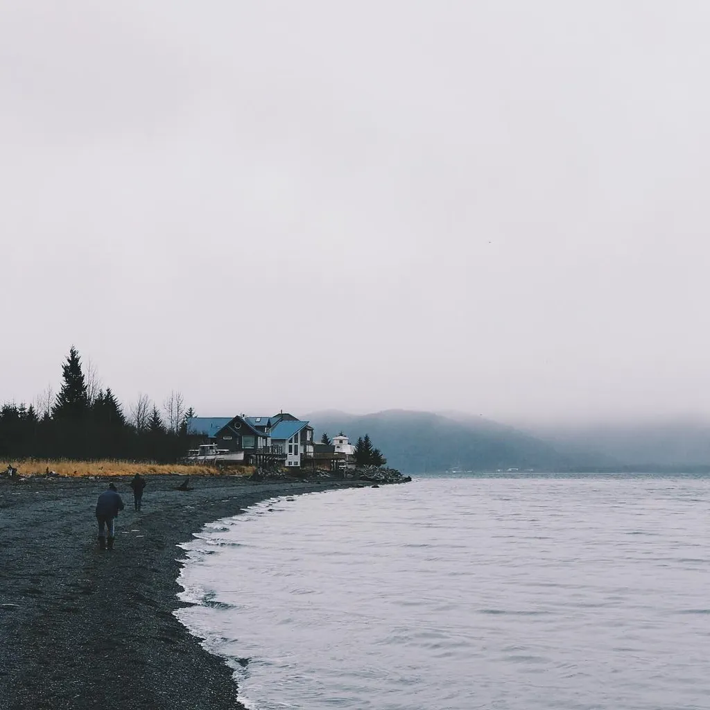 A dark pebble beach meets calm, gray water under a vast, white sky; several buildings sit on the distant shoreline beneath hazy hills.