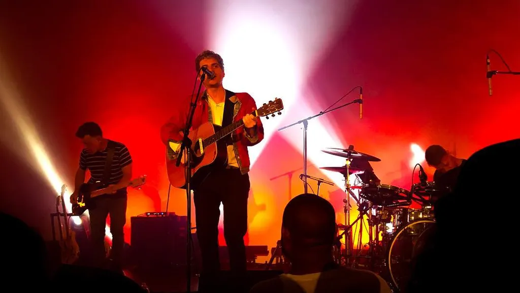 A male musician with a guitar stands center stage, bathed in bright white and red light, while other band members and drum kits are visible in the dramatic, colorful background.