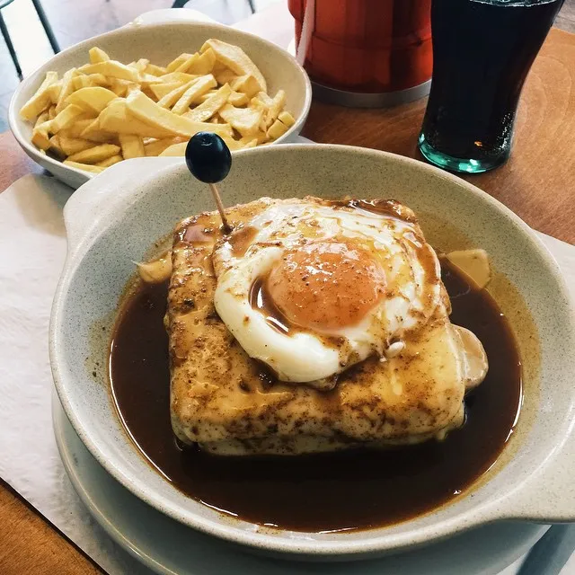 A shallow bowl holds a fried egg and golden bread in dark brown sauce, positioned next to a smaller bowl of crispy yellow potato sticks.