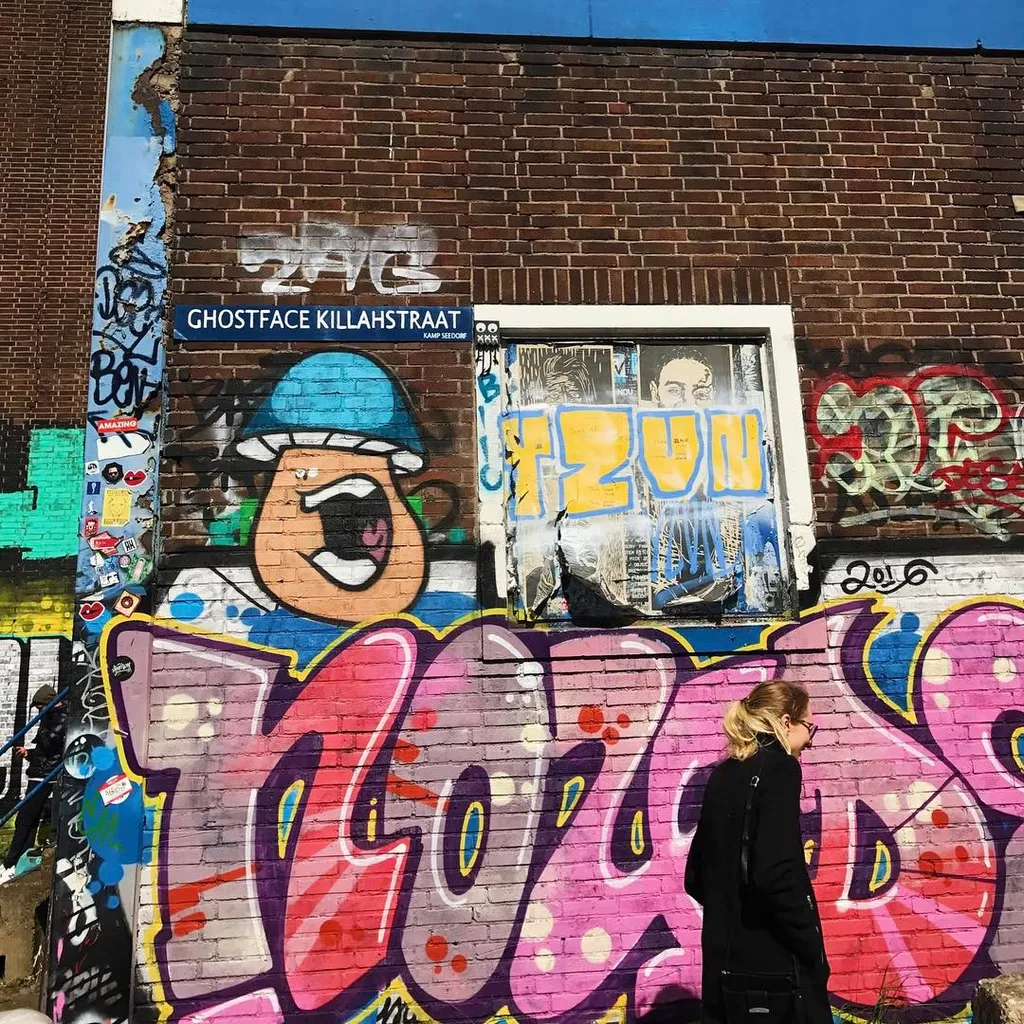 A person stands before a vibrant mural on a brick wall, which displays a cartoon figure, bold lettering, and large, colorful graffiti in shades of pink, purple, and blue.