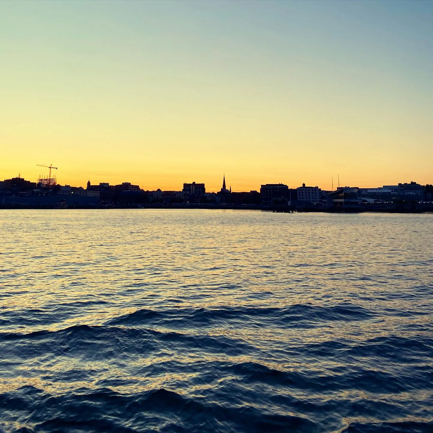 Rippling dark blue water dominates the foreground, meeting a pale yellow and blue sky above a distant, dark silhouette of a city skyline on the horizon.
