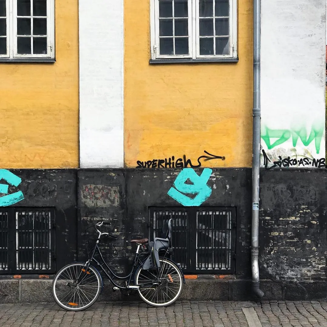 A dark bicycle rests on cobblestones against a bright yellow building wall, which features white trim and windows above a lower, dark section marked by graffiti including teal and white spray paint.