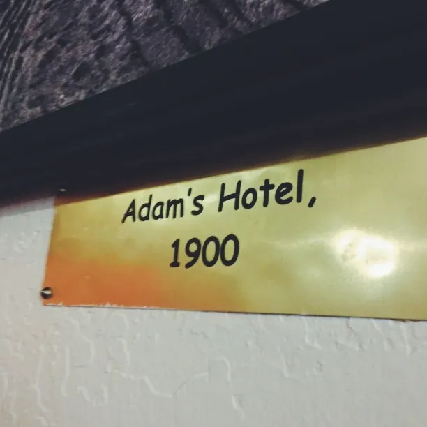 A rectangular sign with a yellow and orange gradient background displays the text Adam's Hotel, 1900, mounted on a textured, light wall.