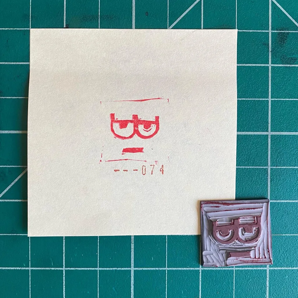 A cream paper on a teal grid displays red pencil markings of a stylized face and the number 31--074, positioned above two small, rectangular, reddish-brown sculpted reliefs, each showing a similar face.