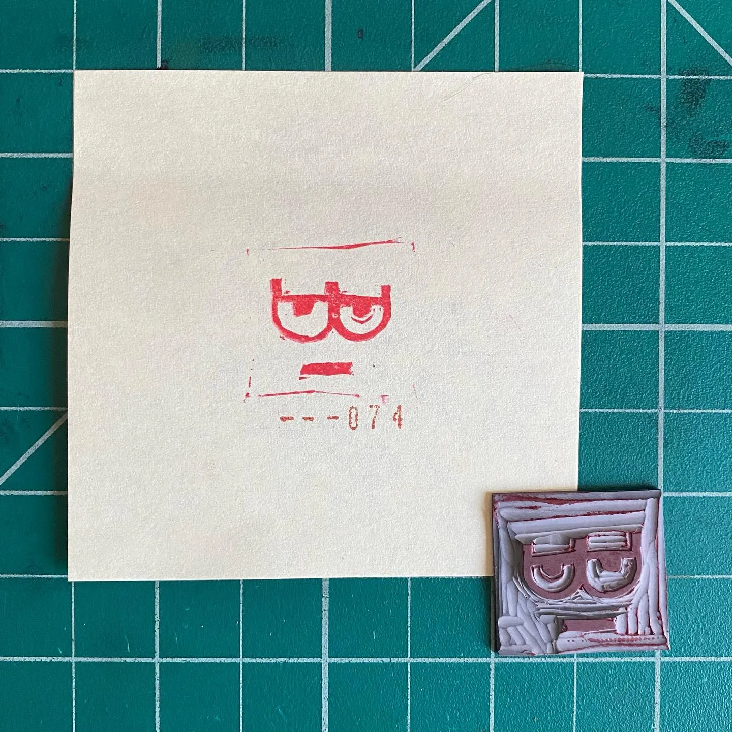 A cream paper on a teal grid displays red pencil markings of a stylized face and the number 31--074, positioned above two small, rectangular, reddish-brown sculpted reliefs, each showing a similar face.