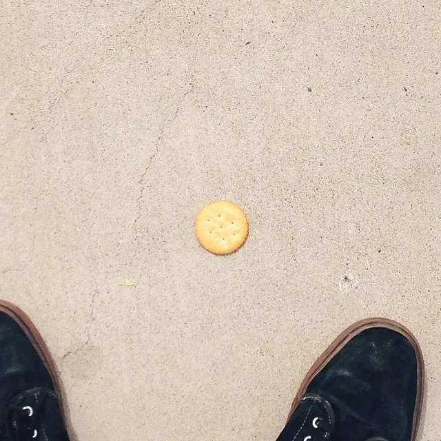 A golden, round cookie sits on textured, light tan sand, framed by a pair of dark, worn shoes at the bottom edge.