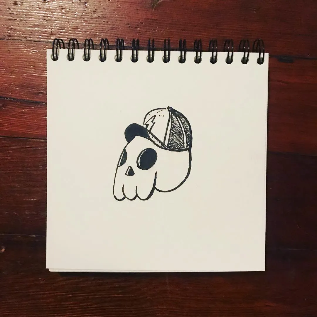 A black ink line drawing of a stylized skull wearing a baseball cap, centered on a spiral-bound sketchbook page resting on a dark wooden surface.