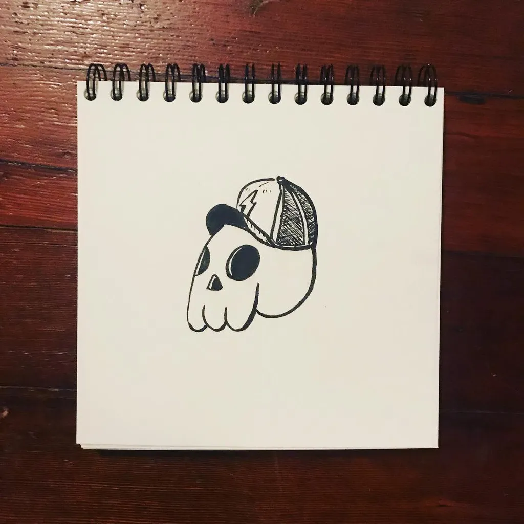 A black ink line drawing of a stylized skull wearing a baseball cap, centered on a spiral-bound sketchbook page resting on a dark wooden surface.