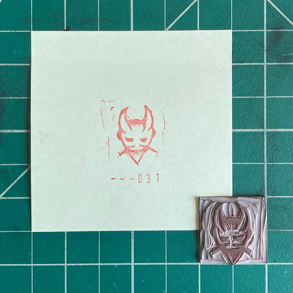 On a teal grid, a cream paper displays a red line drawing of a horned mask and the code --031, next to a small, sculpted pale pink relief of the same mask.