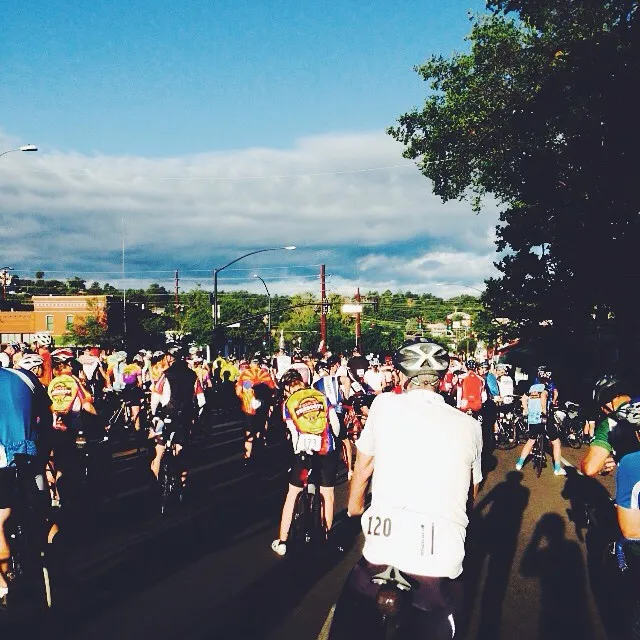 A wide street filled with many people running, framed by lush green trees on the right and a bright blue, cloudy sky above.
