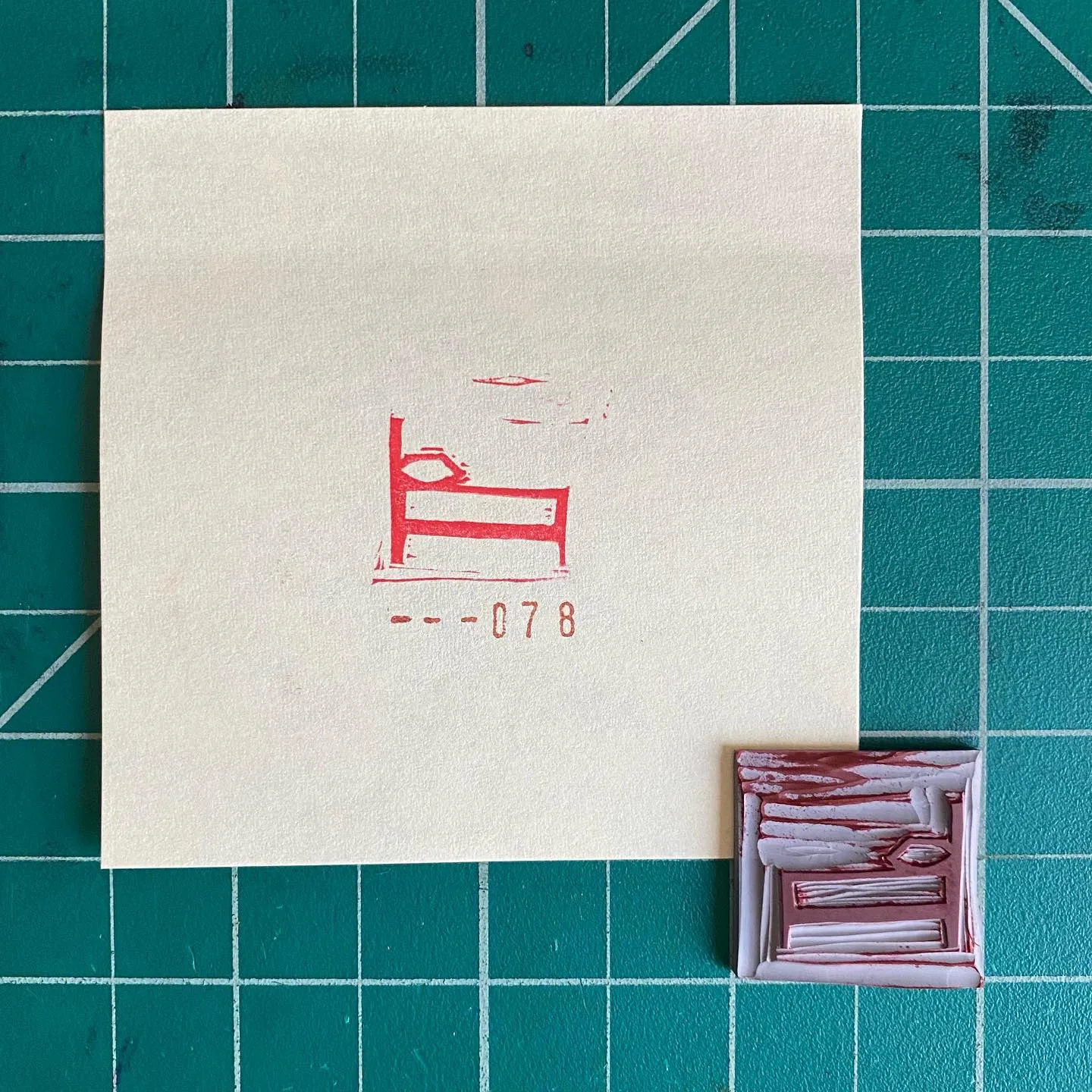 A cream paper on a teal grid displays a red drawing of a bed and the number --078, next to a smaller, square piece featuring layered red and white rectangular marks.