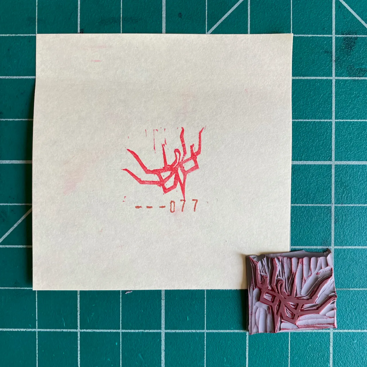 On a teal grid, a cream paper displays a red stylized symbol and the text --077, positioned next to a small, rectangular object featuring layered pink and white abstract patterns.