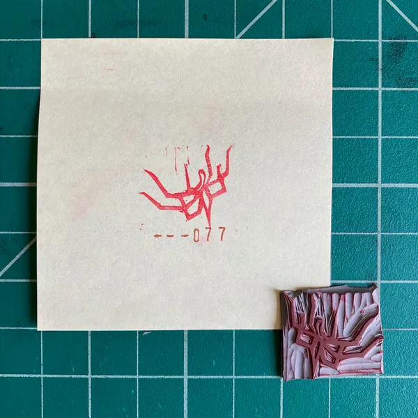 On a teal grid, a cream paper displays a red stylized symbol and the text --077, positioned next to a small, rectangular object featuring layered pink and white abstract patterns.