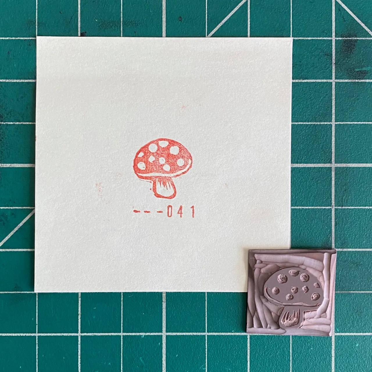 A white card on a green grid background displays a red and white spotted mushroom drawing and the number 041, next to a small, textured, pink square featuring an embossed mushroom design.