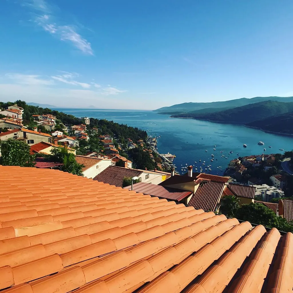 Terracotta roof tiles dominate the foreground, sloping toward a view of a densely built coastal town, a calm blue bay dotted with boats, and hazy blue mountains under a bright sky.