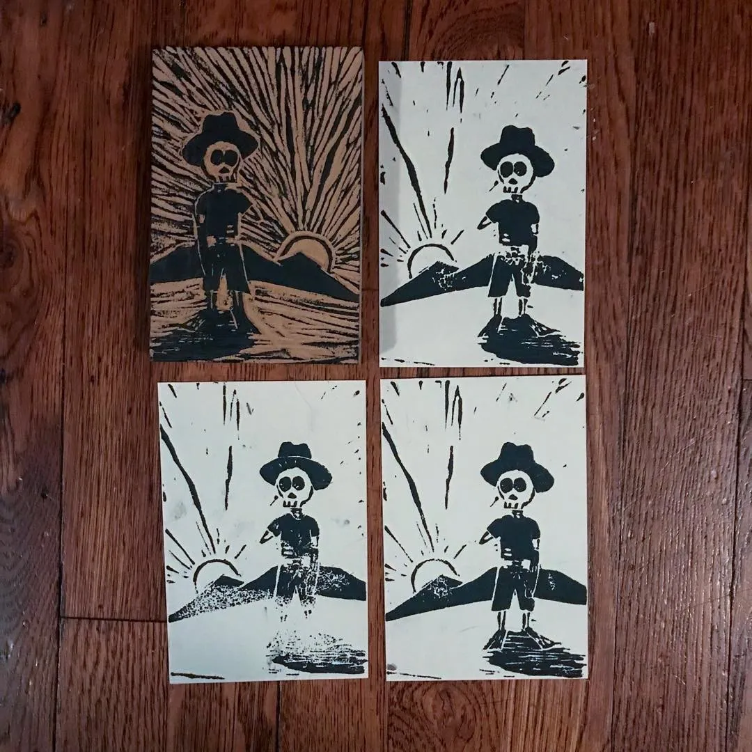 Four square, high-contrast prints are arranged on a wooden floor, depicting a stylized skeleton in a hat against a desert sunset backdrop, featuring distressed black ink marks.