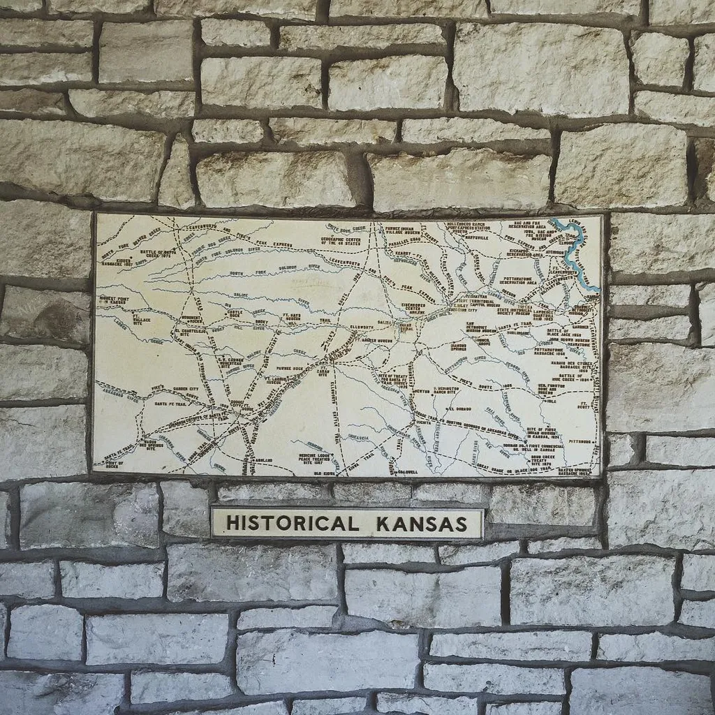 A detailed historical map of Kansas, featuring black lines and text, is affixed to a background of rough, stacked gray stones, with a matching plaque below.