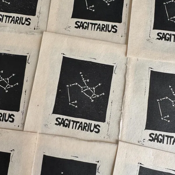 A collection of small, cream-colored prints featuring the black text SAGITTARIUS and a simple black constellation diagram of stars, arranged in a scattered, overlapping pattern.