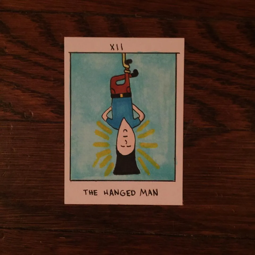 A stylized card depicts a figure hanging upside down from a rope against a light blue background, framed by a yellow sunburst, with the number XII and the title THE HANGED MAN printed below.