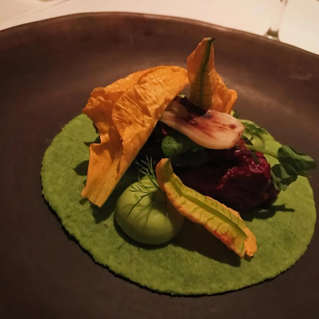 On a dark plate, a bright green circular base supports a dark red mound, surrounded by golden crisps, a small green sphere, and garnishes of pale pink and yellow-green.